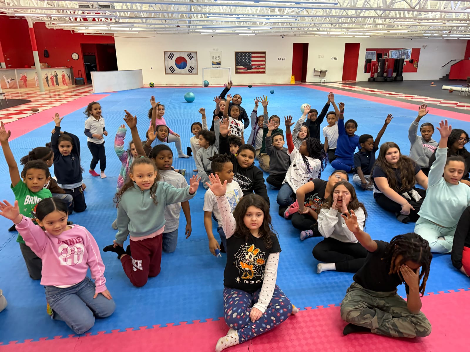 Students kneeling on the mats cheering with hands raised, Korean and American flags on the wall